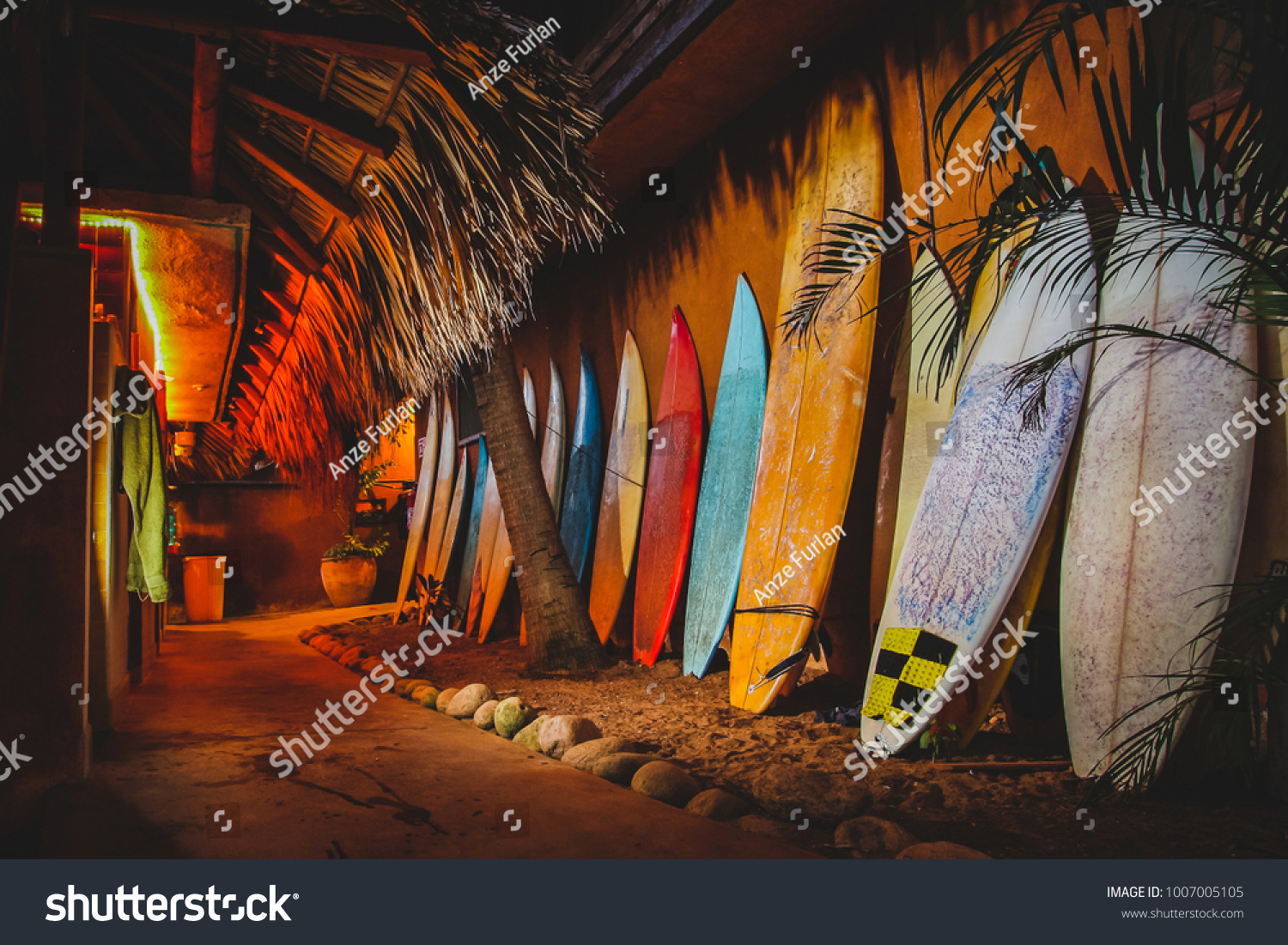 A collection of different colorful surf boards used as a decoration next to a walkway  resting on a wall. Photo taken during the night with a bunch of surfboards on the sand.