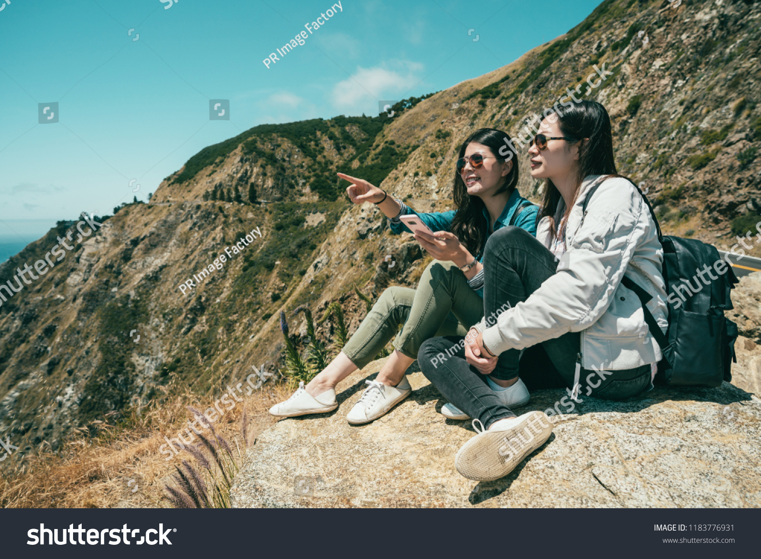 two female tourists finding something by the phone and one girl is pointing somewhere ahead while sitting on the cliff.
