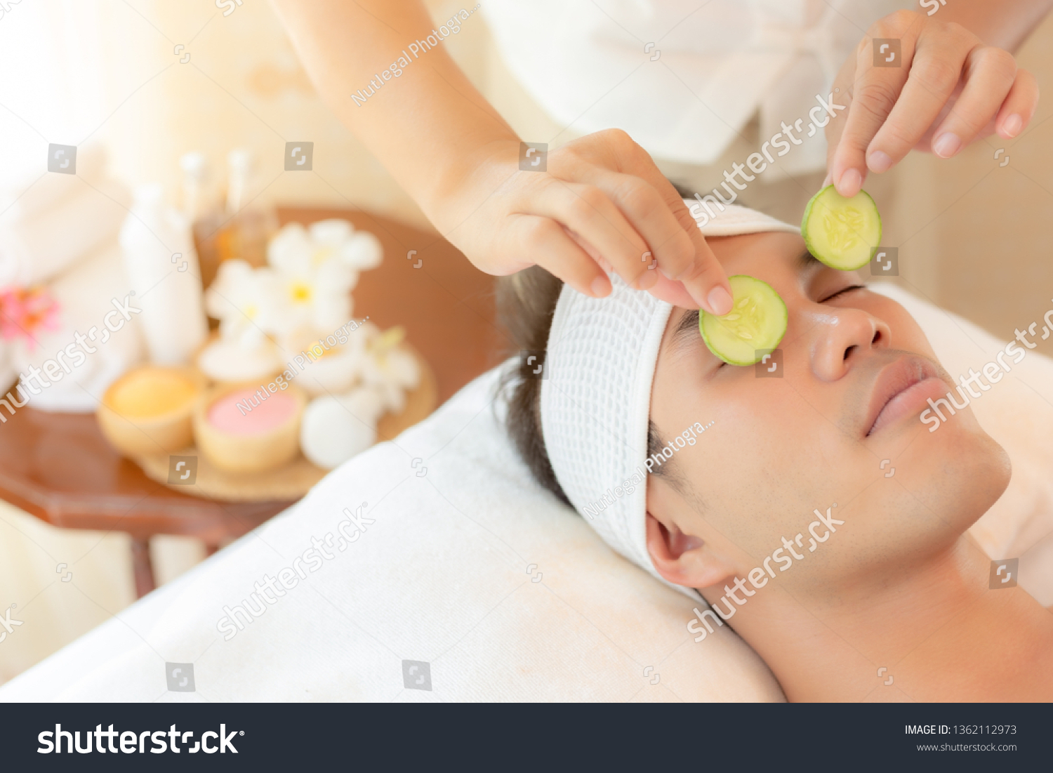 Massage therapist using cucumber slices put to handsome young asian man eyes for restore moisture of eyes skin and solve dark circles under eyes. Attractive handsome young guy use service at spa salon