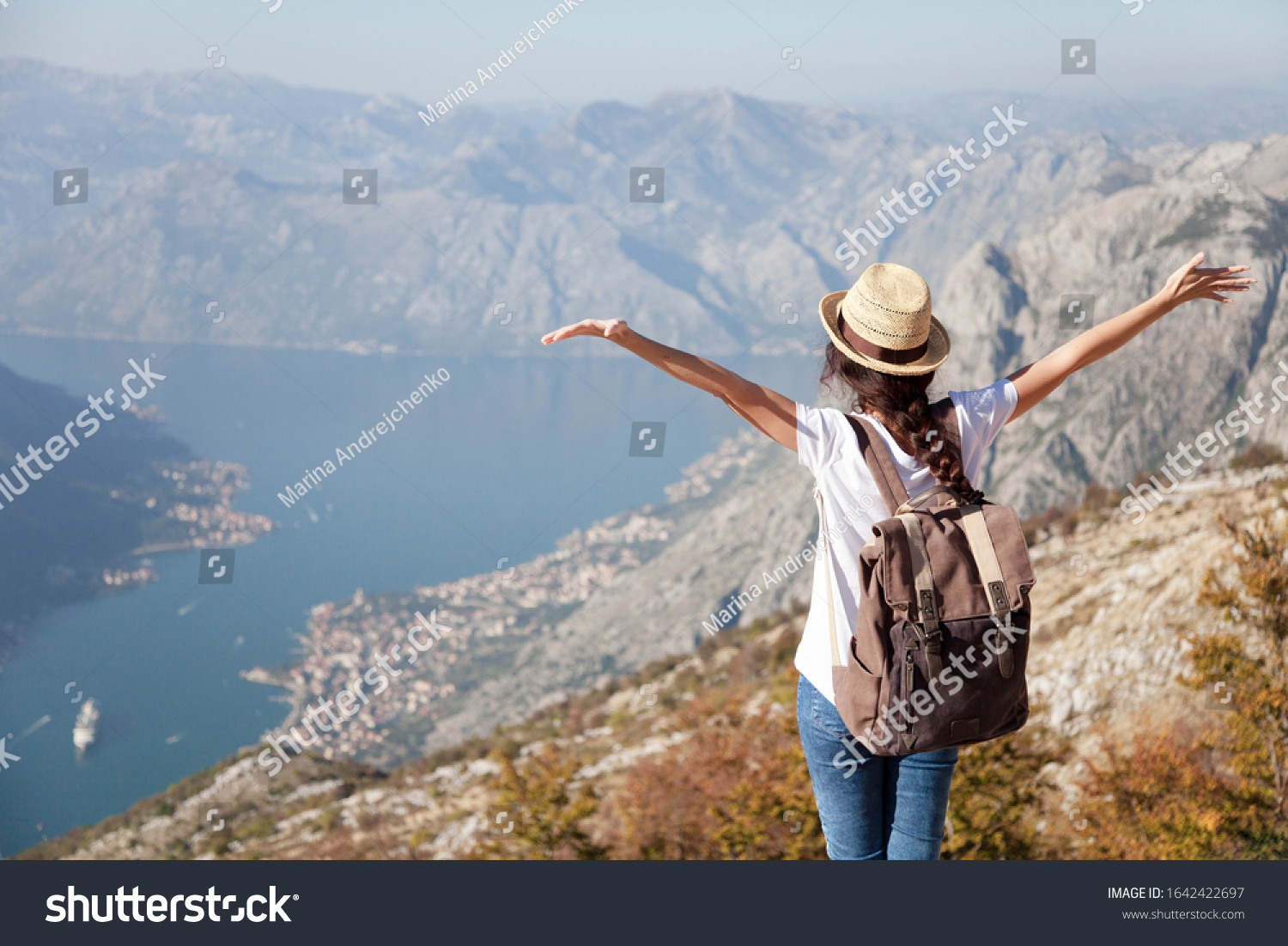 Traveler with backpack on mountain top. Happy woman with raised arms in amazing landscape. Girl tourist enjoying travel adventure freedom vacation. Concept of solo female tourism trip. Rear view.