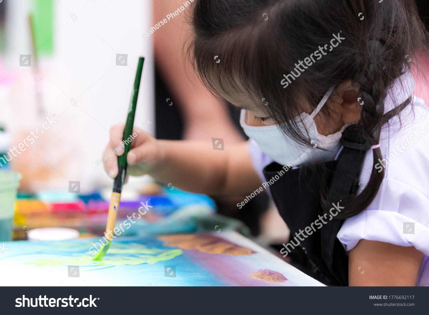 Pupils girl are concentrating on drawing and painting with brush and watercolor on the canvas. An Asian little child is wearing a white cloth face mask while learning to paint. Child 3 years old.