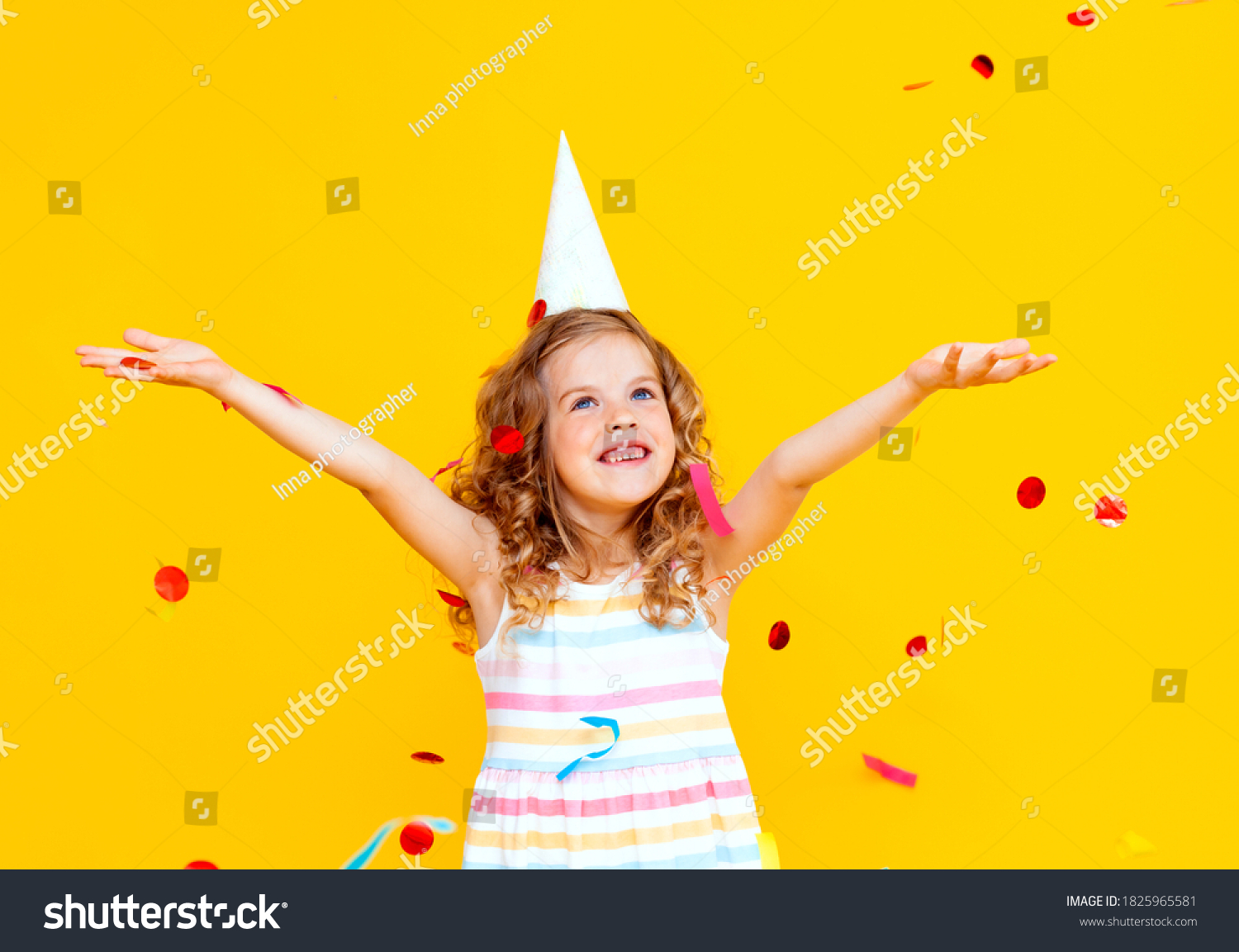 Portrait of a little beautiful girl celebrating her birthday. The kid is throwing colorful confetti. Yellow studio background. The child is having fun