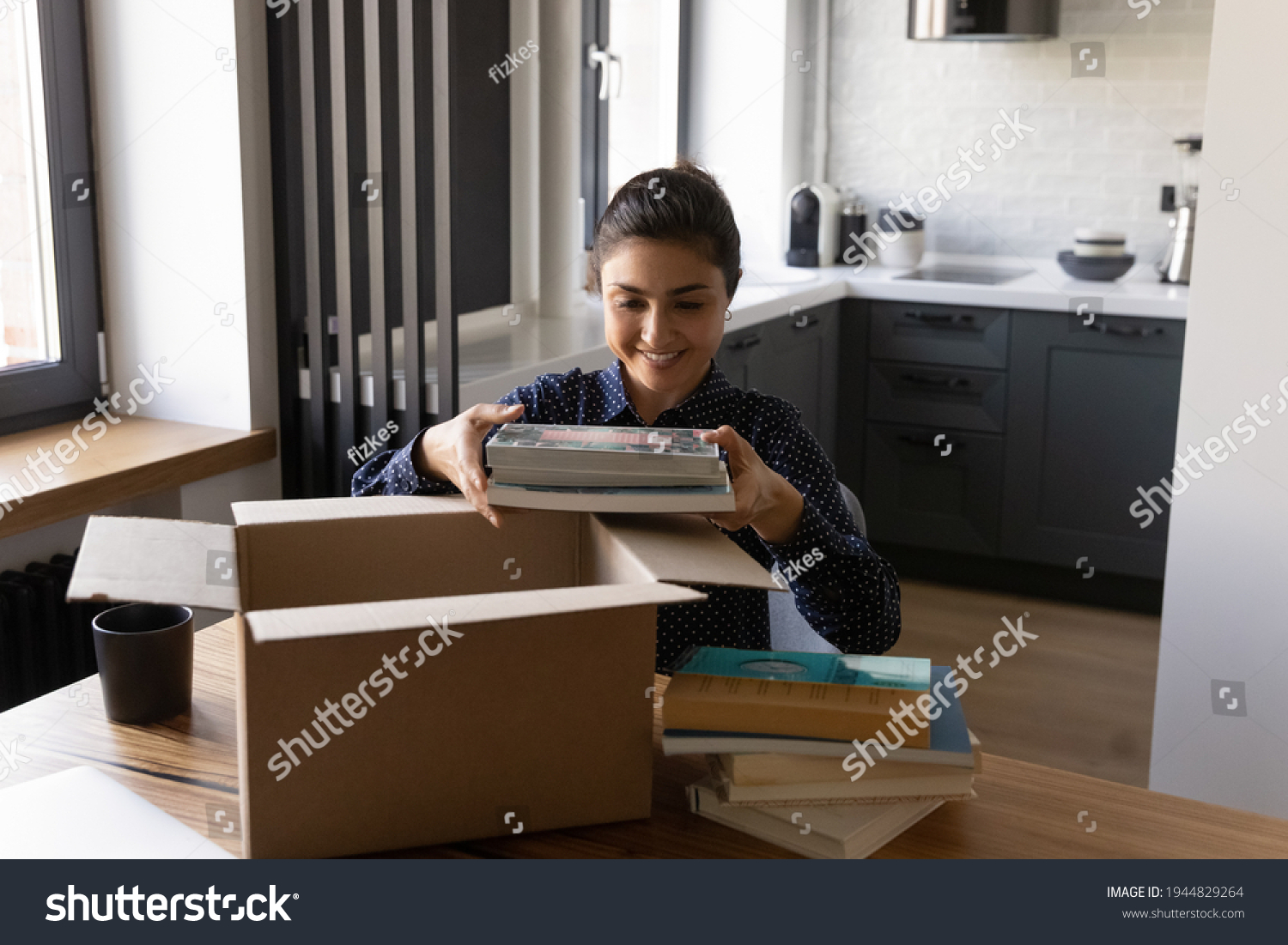 Smiling Indian woman unpacking parcel holding books sitting at table satisfied customer received online store order delivery service happy female packing belongings preparing for relocation