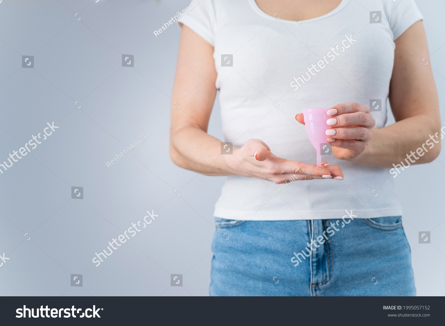 A faceless woman wearing jeans and a white T-shirt holds a pink menstrual cup on a white background. Great alternative to tompons and pads