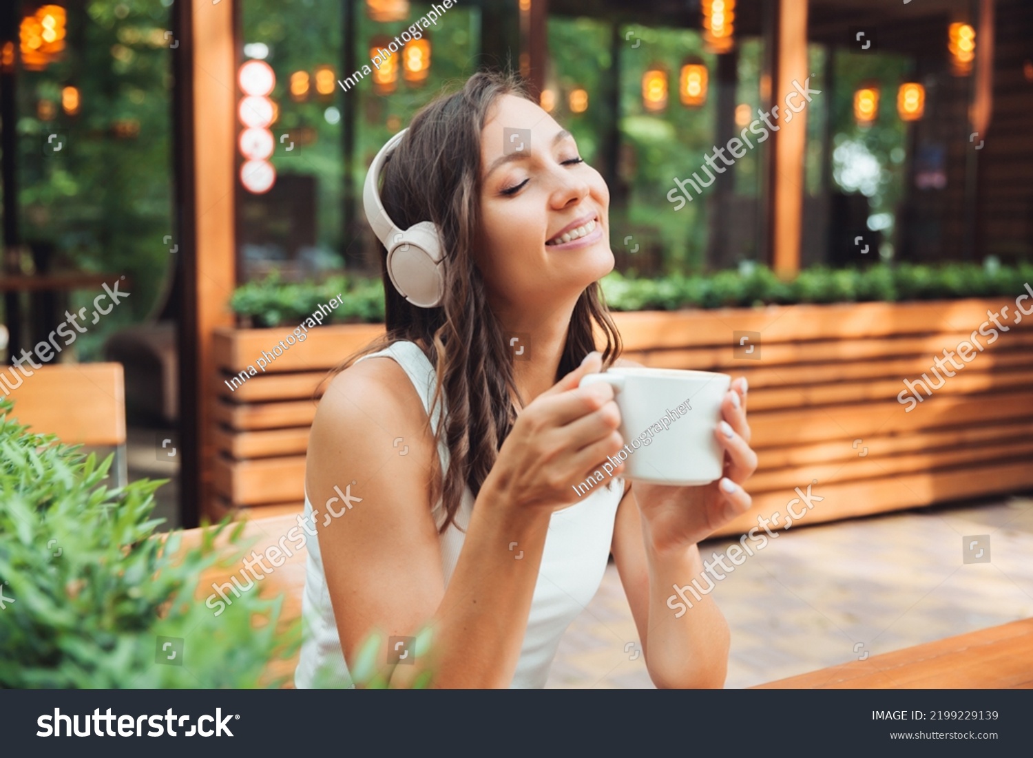 A young beautiful woman with headphones is sitting at a table in a summer cafe and drinking coffee or tea. generation z.