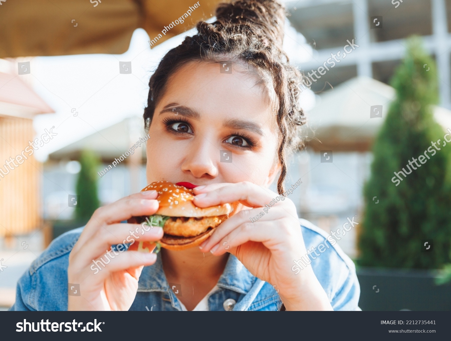 A young glamorous woman with dreadlocks and red lipstick is sitting and eating a burger in a street cafe the concept of eating. long-lasting lipstick