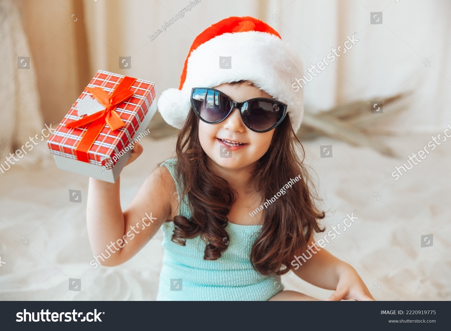 a little girl is sitting on the sand in a Santa hat. the child smiles and looks at the gift box. Merry Christmas. Winter holidays. Space for copying.