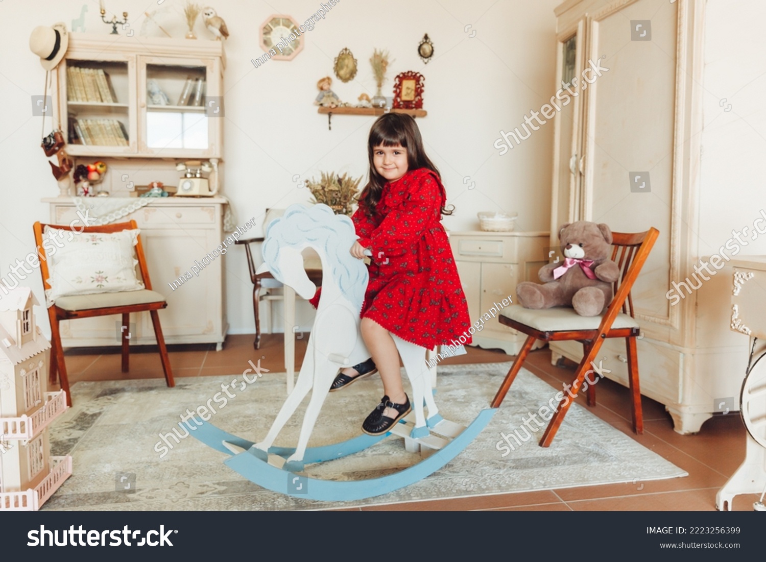 A charming little girl in a dress made of natural material rides a rocking horse in her room. antique nursery interior