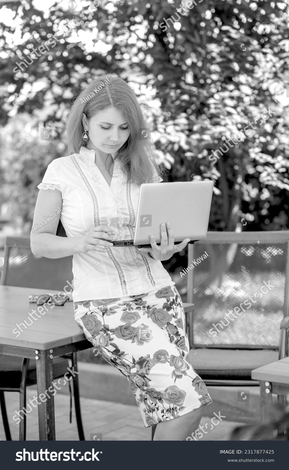 Black and white photo of a young woman working with a laptop in a summer cafe on the street. Coworking. Work outdoors. business concept
