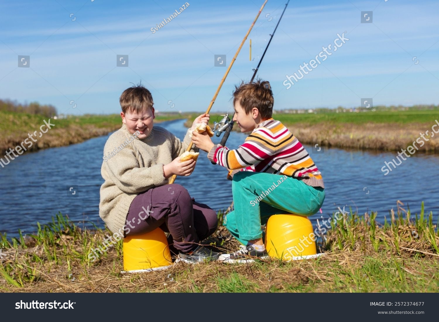 The boys are friends on a fishing trip by the river eating a bun and laughing. Children are friends on a summer fishing trip.
