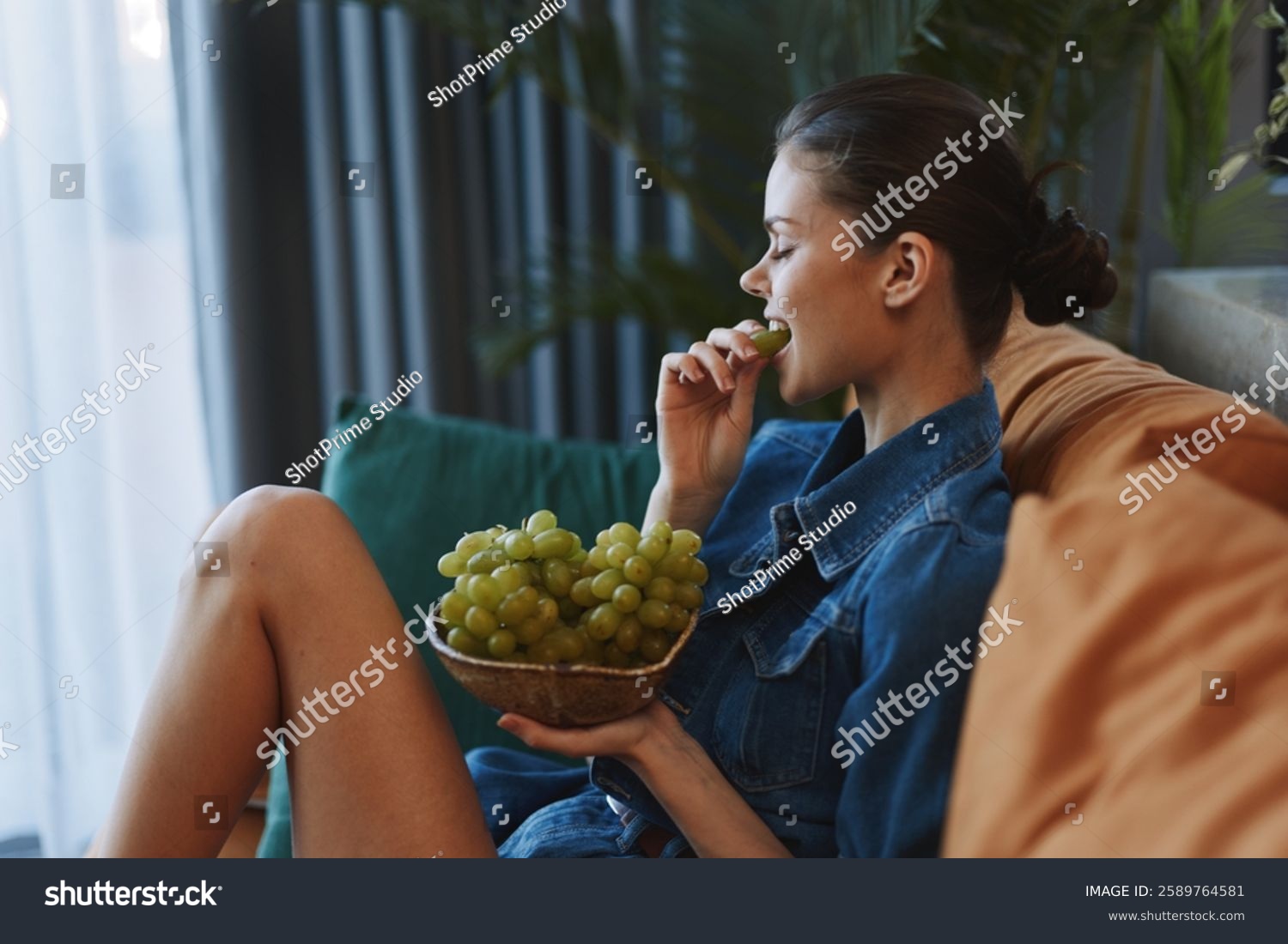 Woman relaxing on a couch with a bowl of grapes in front of her face in a cozy indoor setting