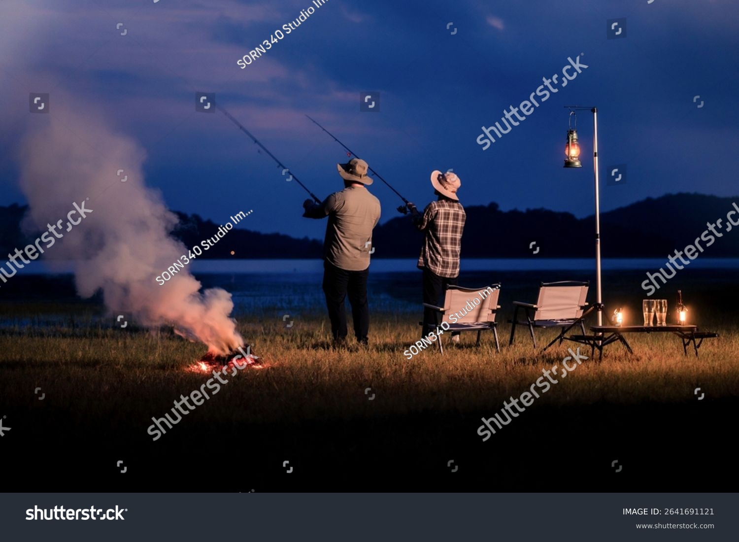 Night fishing with friends by lake under lantern light camping and fishing near campfire at twilight under blue sky Scenic night escape with fishing gear and fire Weekend lakeside activity