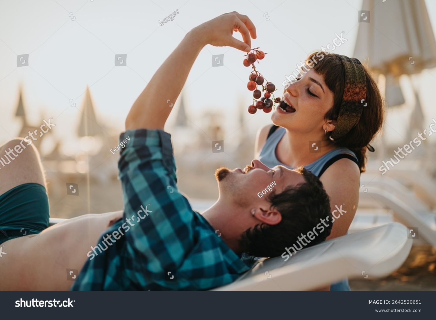 A young couple enjoying their vacation at a sunny beach sharing grapes and relaxing on sun loungers with a joyful atmosphere under umbrellas.