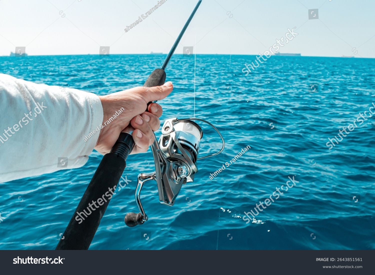 A fisherman hand firmly grips a fishing rod with a modern silver spinning reel ready to cast into the bright blue sea where distant cargo ships dot the horizon
