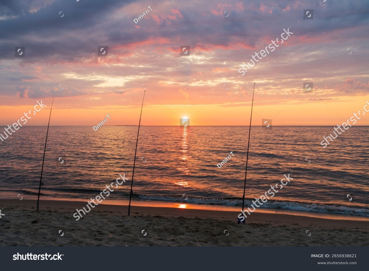 Three fishing rods stand on a sandy beach silhouetted against a breathtaking and colorful sunrise over the tranquil ocean.