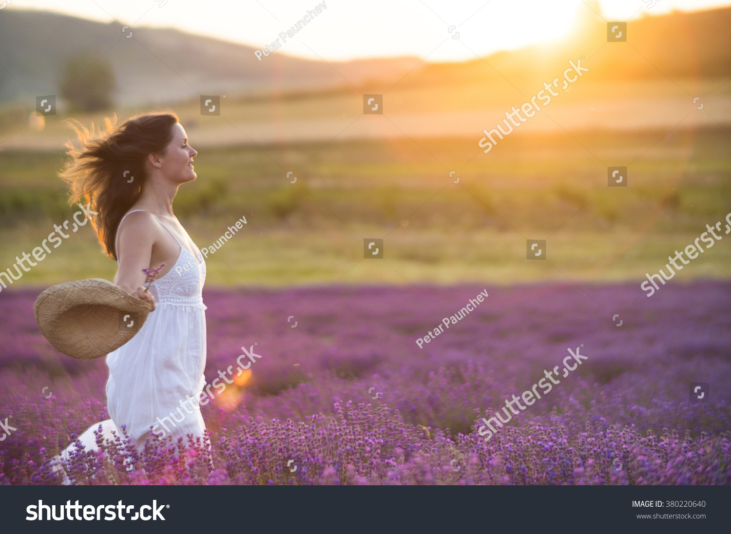 Beautiful young healthy woman with a white dress running joyfully through a lavender field holding a straw hat under the rays of the setting sun.
