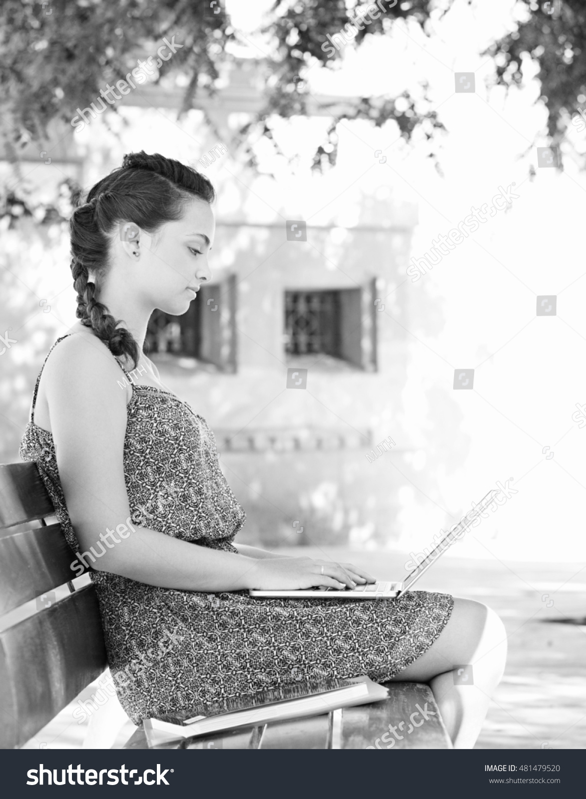 Black and white view of a hispanic teenager student girl sitting on bench in college campus using a laptop computer adolescent technology lifestyle. Young woman studying sunny exterior.