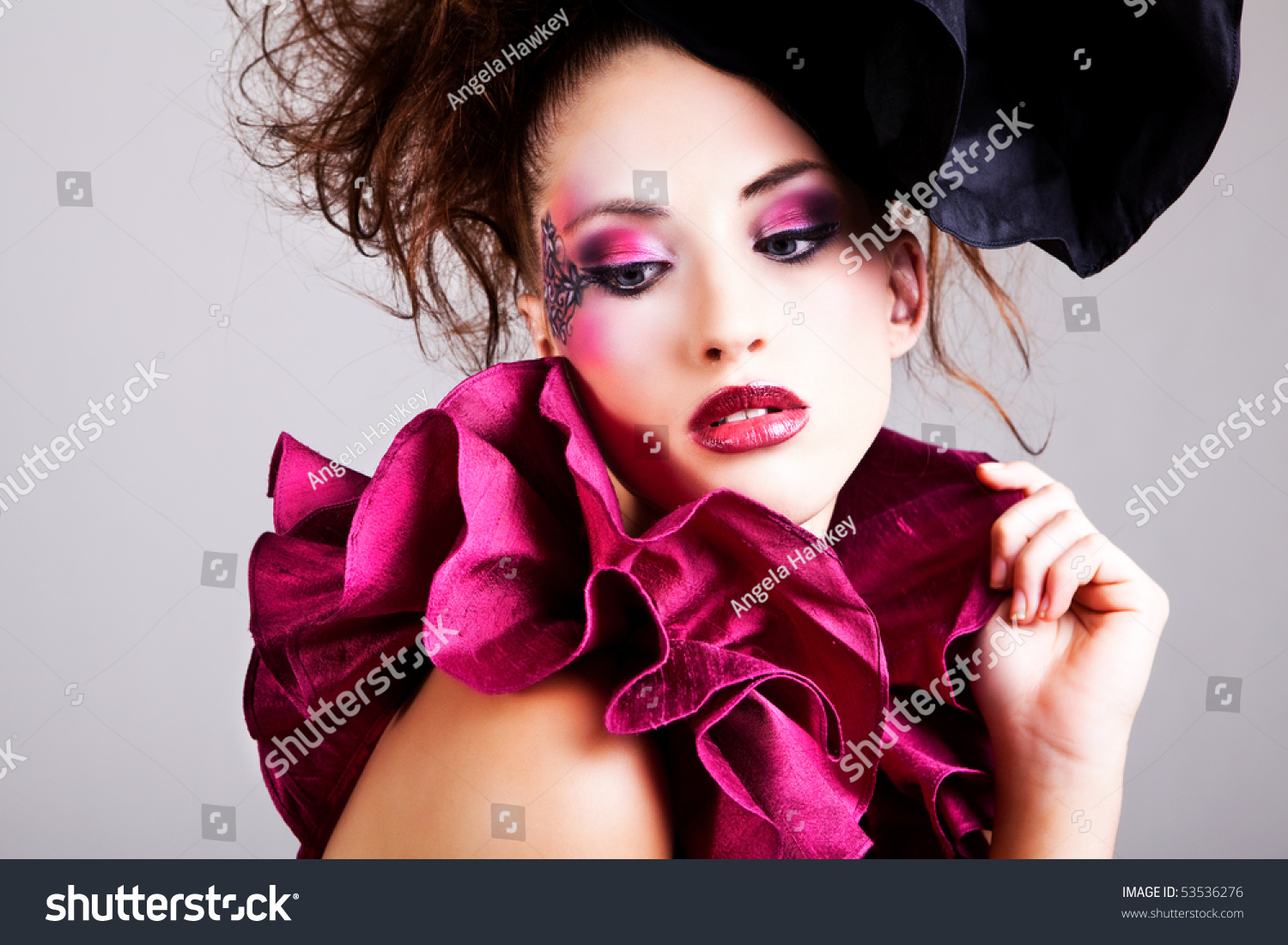 A young woman dressed in avant garde attire. She is wearing a hat and has cosmetic artwork on her right temple. Horizontal shot.
