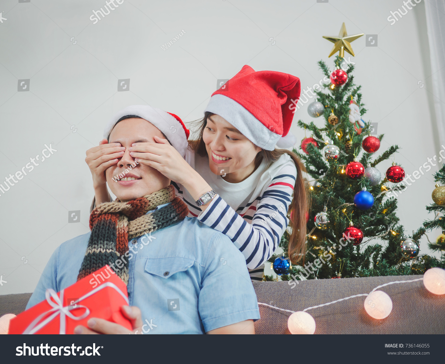 Asian young woman giving a Christmas present to his boyfriend on sofa at home Have Christmas Tree Ornaments.